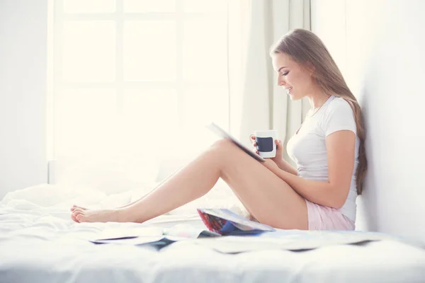 Jeune femme détendue assise sur le lit avec une tasse de café et une tablette numérique — Photo