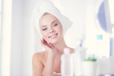 Bornozlu genç kadın banyo aynasına bakıyor.