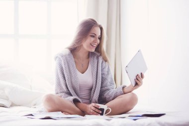 Bir fincan kahve ve dijital tabletle yatakta oturan rahat genç bir kadın.