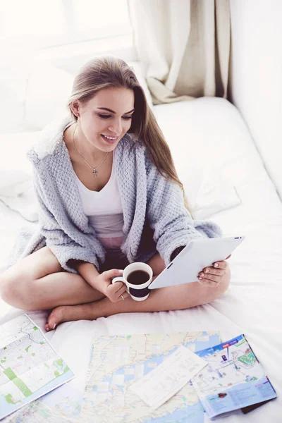 Mujer joven y relajada sentada en la cama con una taza de café y una tableta digital —  Fotos de Stock