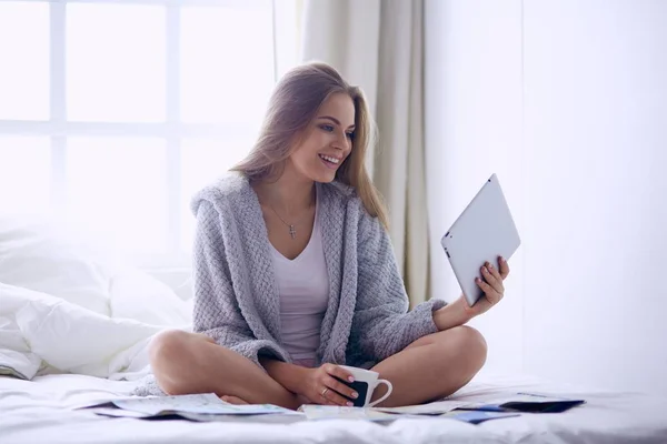 Avslappnad ung kvinna sitter på sängen med en kopp kaffe och digital tablett — Stockfoto