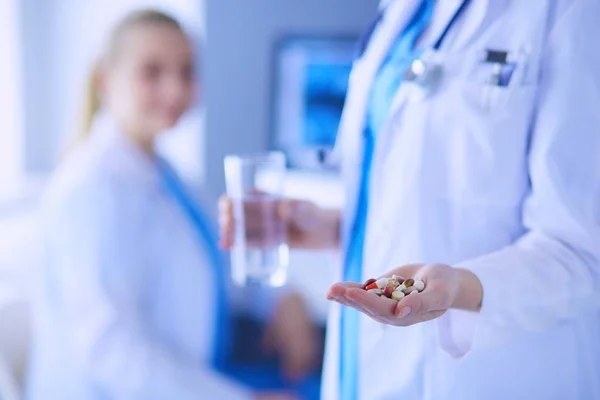 Primer plano tiro de los médicos manos sosteniendo pastillas y vaso de agua en la clínica con su colega en el fondo . —  Fotos de Stock