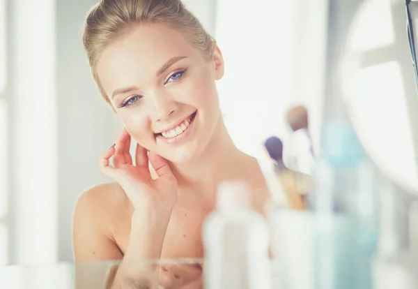 Gyönyörű fiatal nő megérinti az arcát és mosolyog, miközben a tükörbe néz. — Stock Fotó