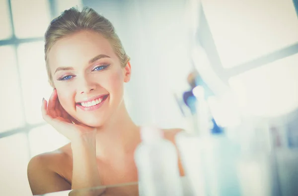 Gyönyörű fiatal nő megérinti az arcát és mosolyog, miközben a tükörbe néz. — Stock Fotó