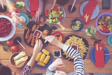 Ahşap masada otururken birlikte yemek yiyen bir grup insanın üst düzey görüntüsü. Masada yemek var. İnsanlar fast food yiyor..