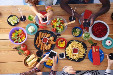 Ahşap masada otururken birlikte yemek yiyen bir grup insanın üst düzey görüntüsü. Masada yemek var. İnsanlar fast food yiyor..