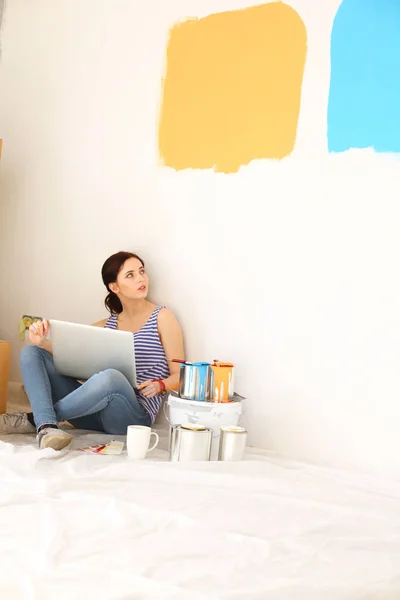 Retrato de mujer joven mientras pinta nuevo apartamento, sentado con el ordenador portátil. Mujer joven con portátil —  Fotos de Stock