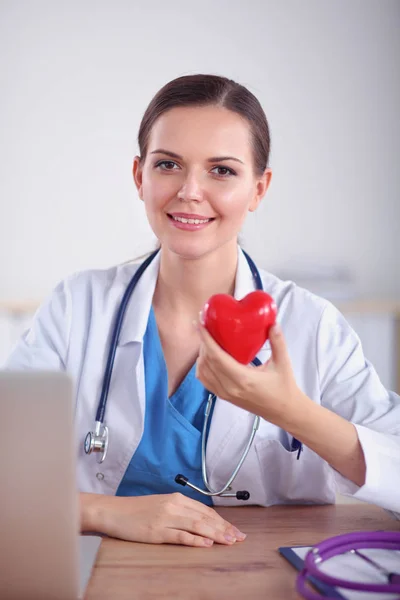 Vackra unga leende kvinnliga läkare sitter vid skrivbordet och hålla hjärtat. — Stockfoto