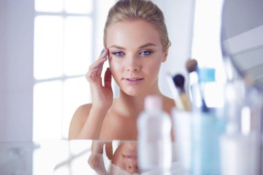 Bornozlu genç kadın banyo aynasına bakıyor.