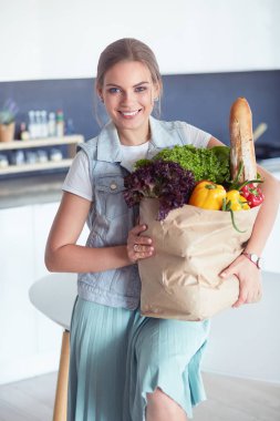 Sebzeli alışveriş poşeti taşıyan genç bir kadın. Mutfakta duruyor.