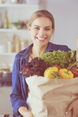 Sebzeli alışveriş poşeti taşıyan genç bir kadın. Mutfakta duruyor.