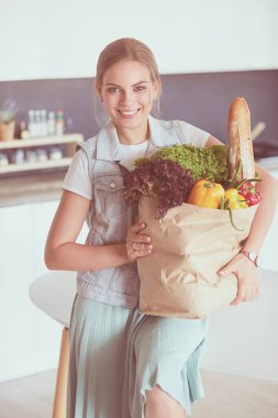 Sebzeli alışveriş poşeti taşıyan genç bir kadın. Mutfakta duruyor.