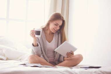 Bir fincan kahve ve dijital tabletle yatakta oturan rahat genç bir kadın.