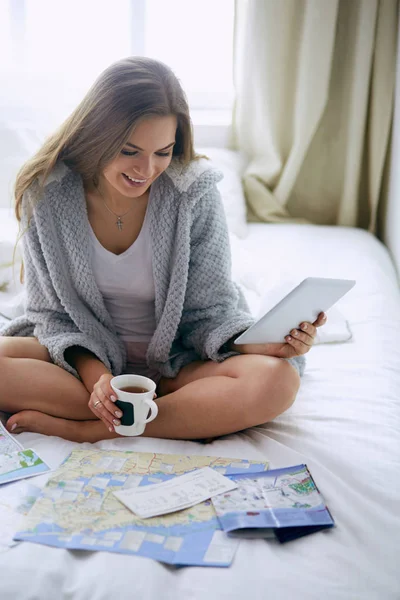 Mujer joven y relajada sentada en la cama con una taza de café y una tableta digital —  Fotos de Stock