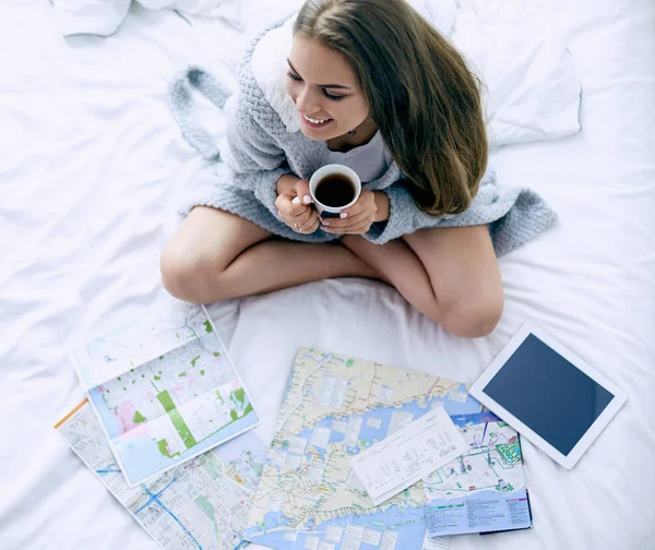 Avslappnad ung kvinna sitter på sängen med en kopp kaffe och digital tablett — Stockfoto