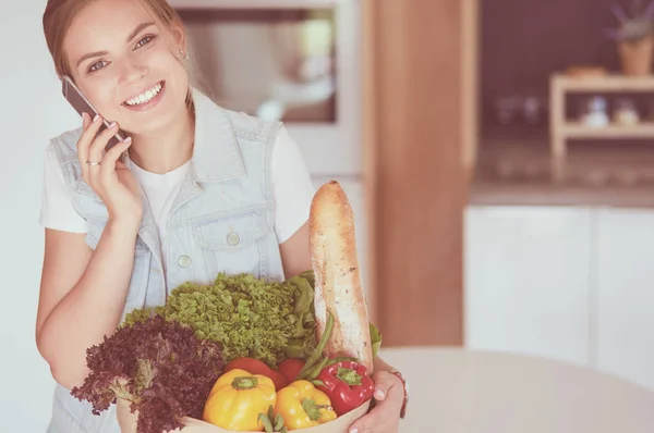 Leende kvinna med mobiltelefon håller shoppingväska i köket — Stockfoto