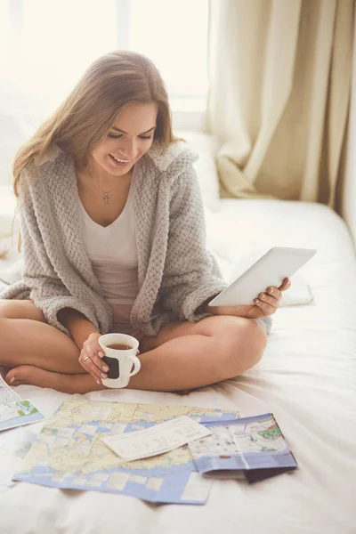 Mujer joven y relajada sentada en la cama con una taza de café y una tableta digital —  Fotos de Stock