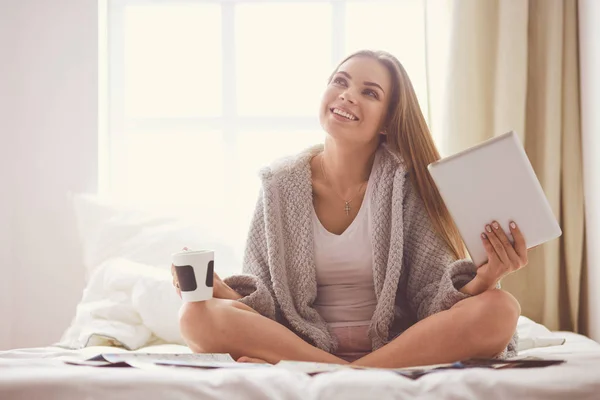 Relaxed young woman sitting on bed with a cup of coffee and digital tablet — Stock Photo, Image