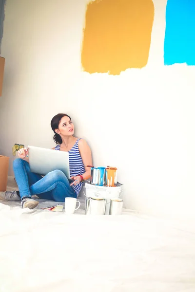 Retrato de mujer joven mientras pinta nuevo apartamento, sentado con el ordenador portátil. Mujer joven con portátil — Foto de Stock