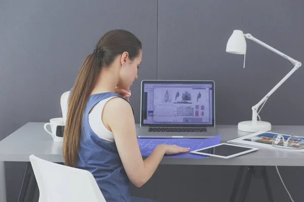Ung kvinde sidder ved skrivebordet med instrumenter, plan og laptop. - Stock-foto
