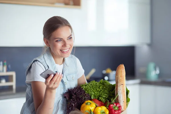 Mujer joven sosteniendo bolsa de la compra de comestibles con verduras. De pie en la cocina —  Fotos de Stock