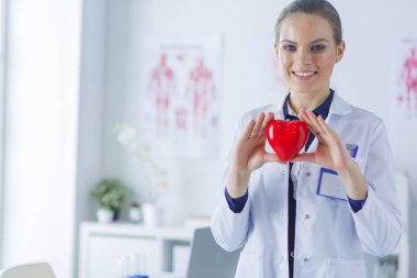 Steteskoplu bir doktor, beyaz arka planda kırmızı kalbi inceliyor.