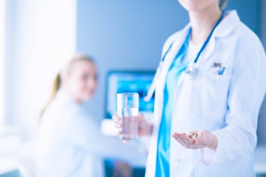 Doktorun arka planda meslektaşıyla klinikte elinde haplar ve bir bardak su tutarken görüntüsü..