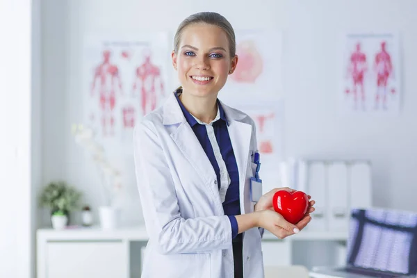 Jonge vrouw arts met een rood hart, staande op ziekenhuis achtergrond — Stockfoto