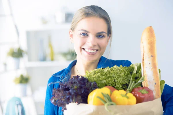 Fiatal nő gazdaság élelmiszer bevásárló táska zöldségekkel.Álló a konyhában — Stock Fotó