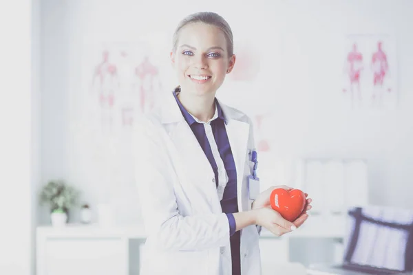 Mladá žena lékař drží červené srdce, stojí na nemocničním pozadí — Stock fotografie