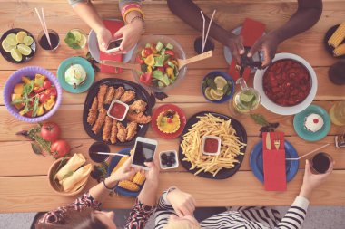 Ahşap masada otururken birlikte yemek yiyen bir grup insanın üst düzey görüntüsü. Masada yemek var. İnsanlar fast food yiyor..