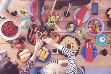 Ahşap masada otururken birlikte yemek yiyen bir grup insanın üst düzey görüntüsü. Masada yemek var. İnsanlar fast food yiyor..