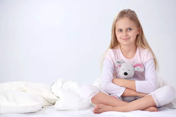 Malá holčička v ložnici sedí na posteli. Holčička je nosí pyžamo a seděl v posteli . — Stock fotografie