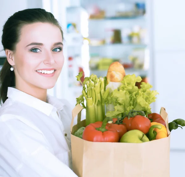 Mujer joven sosteniendo bolsa de la compra de comestibles con verduras.De pie en la cocina. Mujer en la cocina mirando a la cámara — Foto de Stock