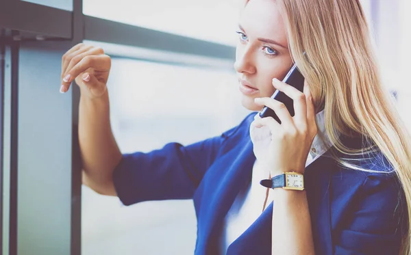 Podnikatelka stojí proti oknu kanceláře a mluví na mobilním telefonu — Stock fotografie