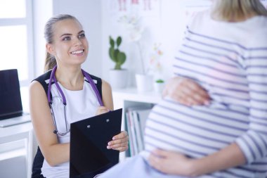 Steteskoplu ve tabletli genç kadın doktor hastanede hamile bir kadınla konuşuyor..