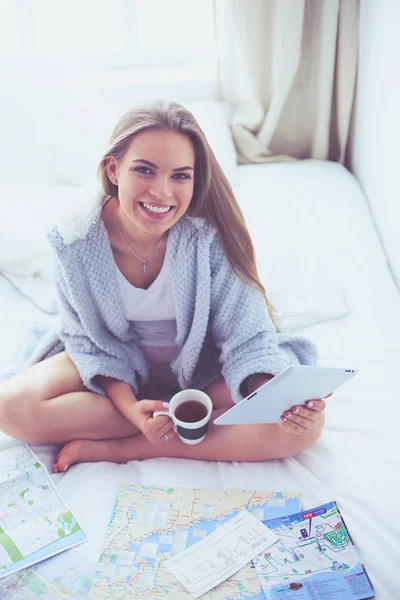 Avslappnad ung kvinna sitter på sängen med en kopp kaffe och digital tablett — Stockfoto