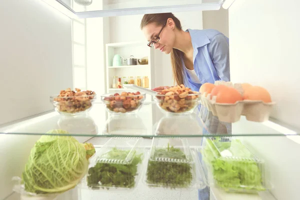 Портрет женщины, стоящей рядом с открытым холодильником, полным здоровой пищи, овощей и фруктов. — стоковое фото