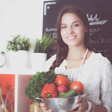Mutfakta elinde sebzelerle gülümseyen genç bir kadın.