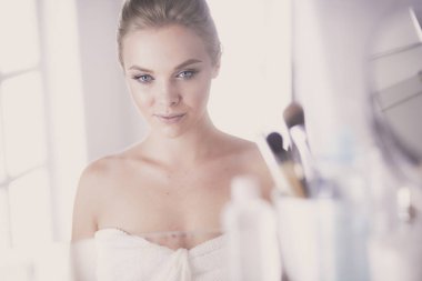 Bornozlu genç kadın banyo aynasına bakıyor.