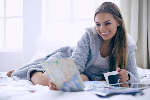 Avslappnad ung kvinna sitter på sängen med en kopp kaffe och digital tablett — Stockfoto