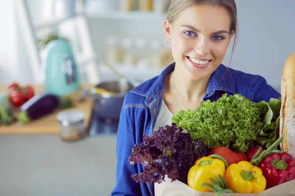 Sebzeli alışveriş poşeti taşıyan genç bir kadın. Mutfakta duruyor.