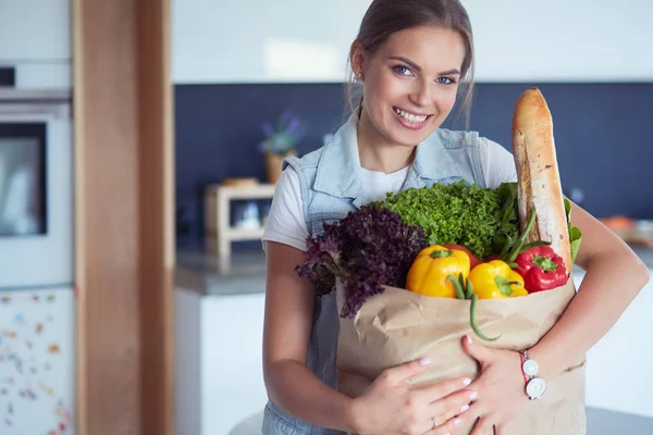 Jeune femme tenant sac d'épicerie avec des légumes. Debout dans la cuisine — Photo