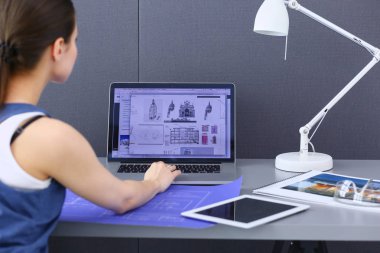 Masasında enstrümanlar, planlar ve dizüstü bilgisayarlarla oturan genç bir kadın..