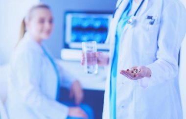 Doktorun arka planda meslektaşıyla klinikte elinde haplar ve bir bardak su tutarken görüntüsü..
