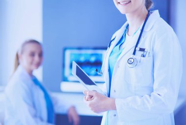 Doktorun arka planda meslektaşıyla klinikte elinde haplar ve bir bardak su tutarken görüntüsü..