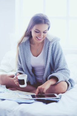 Bir fincan kahve ve dijital tabletle yatakta oturan rahat genç bir kadın.