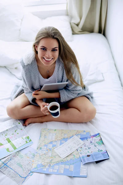 Розслаблена молода жінка сидить на ліжку з чашкою кави та цифровим планшетом — стокове фото