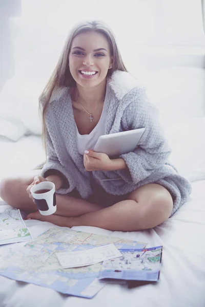 Mujer joven y relajada sentada en la cama con una taza de café y una tableta digital —  Fotos de Stock