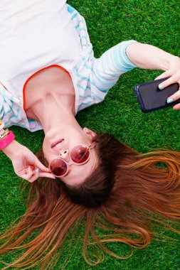Güzel genç bir kadın yeşil çimenlerde yatarken telefonuyla selfie çekiyor. Güzel genç bir kadın selfie çekiyor.